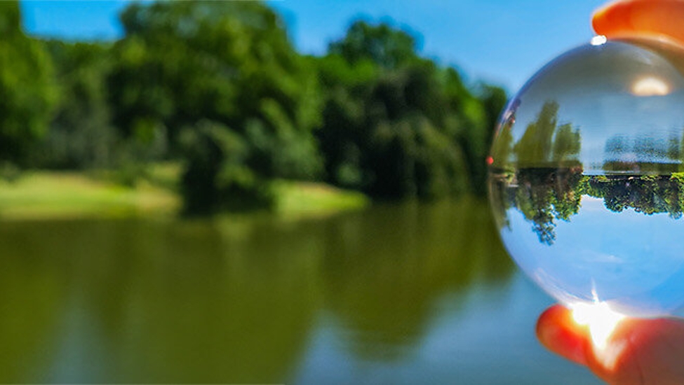 Eine Hand hält eine transparente Glaskugel, durch die ein See und ein umliegender Wald zu sehen sind, die Natur erscheint darin gestochen scharf