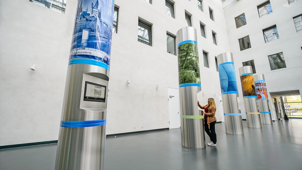 Das Foyer des Verwaltungsgebäudes der StEB Köln. In dem Foyer stehen fünf Metallsäulen. Diese Säulen sind gleichzeitig auch Infostände. An der zweiten Säule von links steht eine Mitarbeiterin, die sich den Infoscreen ansieht.
