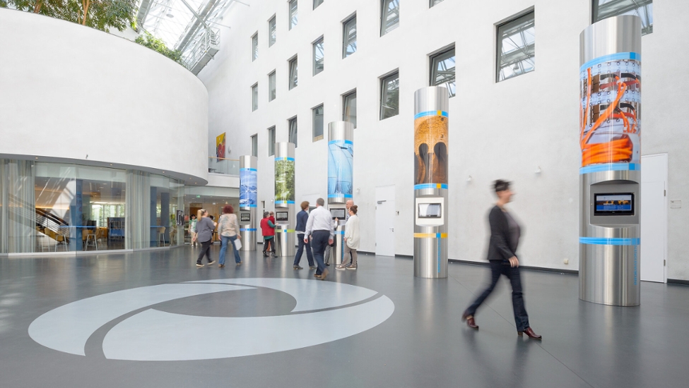 Das Foyer des Verwaltungsgebäudes der StEB Köln. Im Hintergrund stehen fünf Metallsäulen. Im Vordergrund befinden sich Mitarbeitende der StEB Köln, die sich unterhalten und mit dem Rücken zur Kamera stehen. Das Logo der StEB Köln befindet sich auf dem Boden.