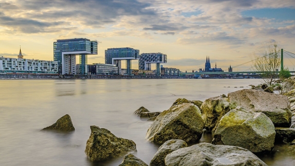Die Kranhäuser am Rhein. Das Foto wurde von der rechten Rheinseite aufgenommen und im unteren Teil des Fotos sind große Steine zu sehen. Es ist Abenddämmerung und ein bewölkter Tag.