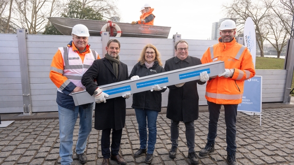 Fünf Personen halten einen Dammbalken einer Hochwasserschutzwand hoch. Im Hintergrund ist ein Teil einer Hochwasserschutzwand aufgebaut.