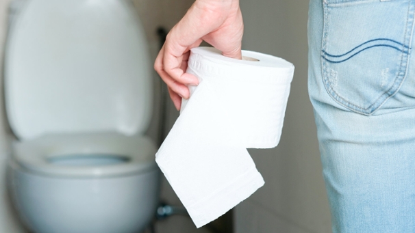 Eine Person steht vor einer Toilette und hält eine Rolle Toilettenpapier in der Hand.