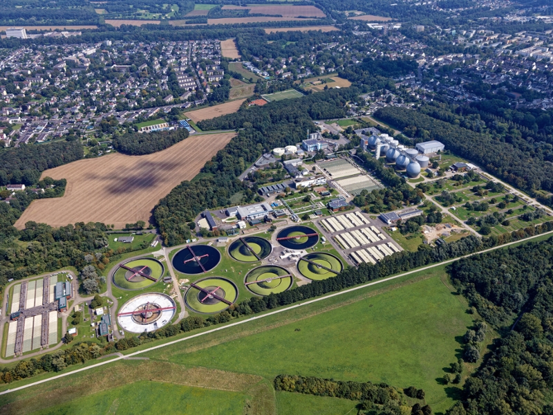 Luftbild eines Klärwerks mit angrenzendem Feld und Wohnsiedlung.